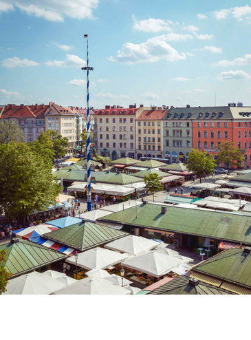 Exterior view LOUIS Hotel Munich Viktualienmarkt View of the colorful Viktualienmarkt in Munich with maypole, surrounded by historic buildings under a blue sky.