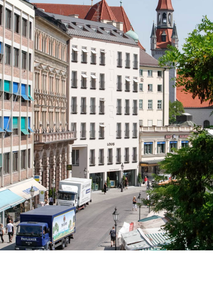 Aussenansicht LOUIS Hotel Muenchen Viktualienmarkt LOUIS Hotel in München, modernes Gebäude in urbaner Umgebung, mit Marktständen und Kirchturm im Hintergrund.
