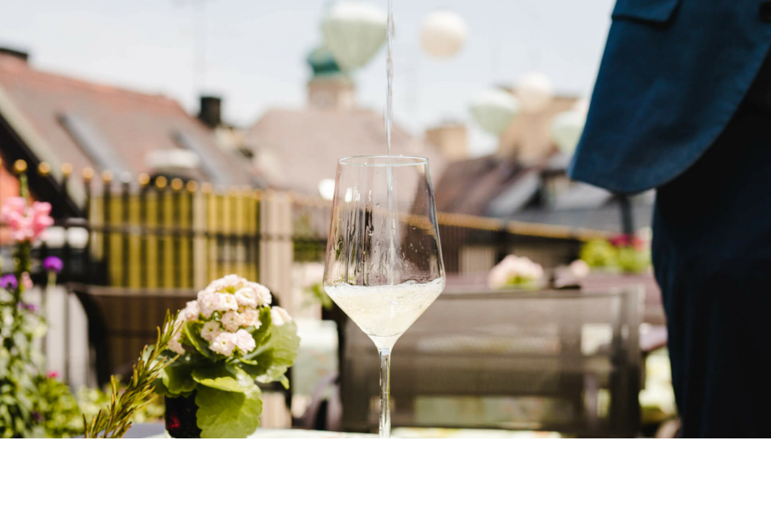 The Roof Terrace | Kühle Drinks auf der Dachterrasse des LOUIS Hotel München Weißwein wird auf einer sonnigen Hotelterrasse in ein Glas eingeschenkt, mit Blumen und Stadtblick im Hintergrund.