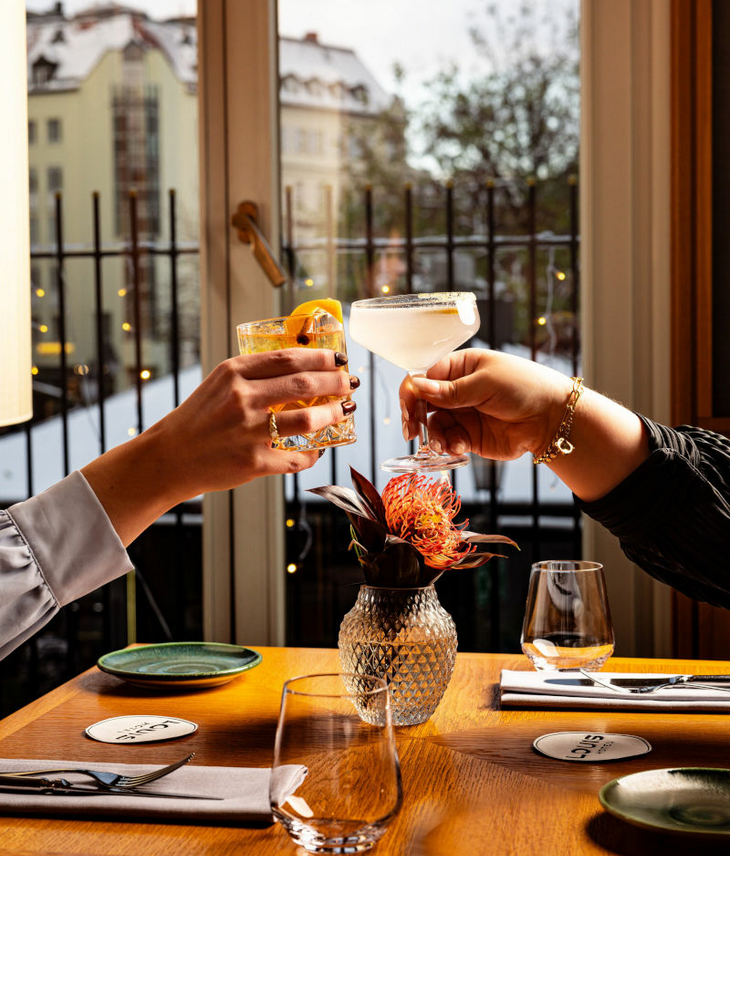 Zwei Hände stoßen mit Getränken über einem gedeckten Restauranttisch im LOUIS Hotel an. Auf dem Tisch stehen Gläser, Teller, Besteck und eine kleine Vase mit einer orangefarbenen Blume. Im Hintergrund ist ein großes Fenster.