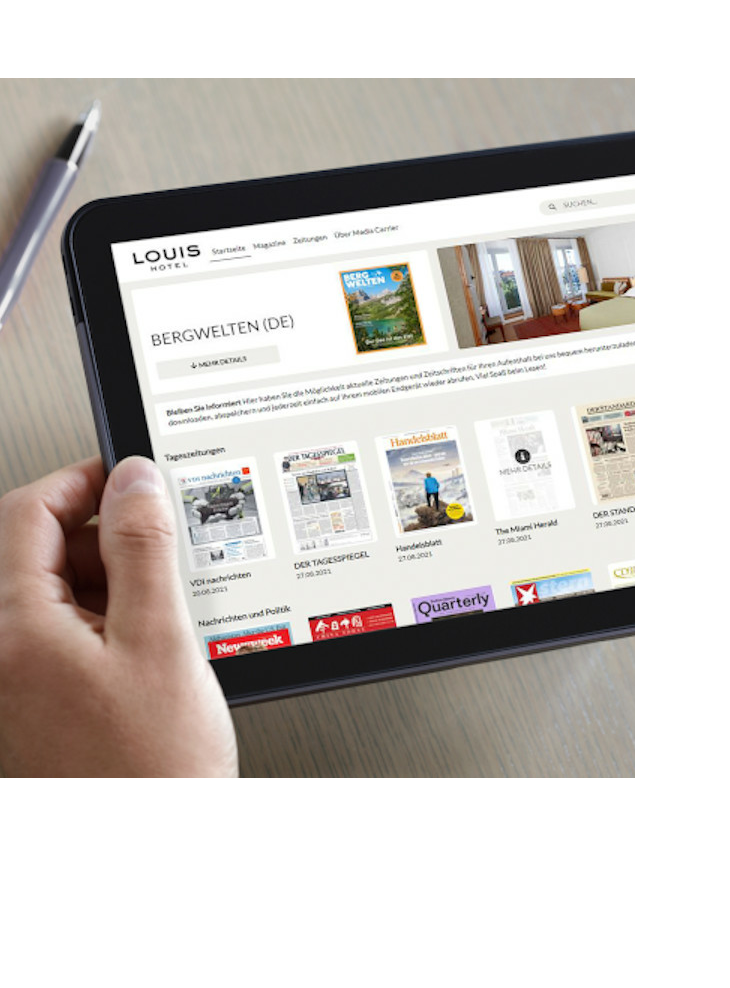 Mediabox - digitale Inhalte l LOUIS Hotel München Person hält ein Tablet mit einer Hotelwebseite, daneben eine Tasse Kaffee auf einem Tisch.