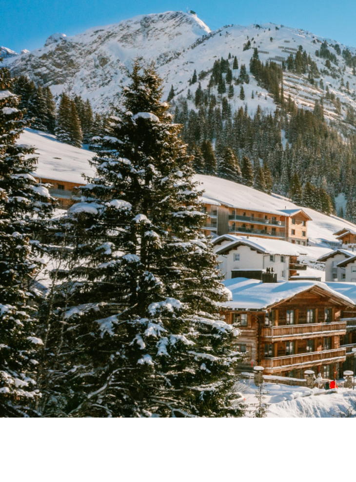 Panoramic view of Lech – The Alpine Retreat Snowy alpine landscape with sun above Hotel Severin*s in Lech, surrounded by mountains and trees.