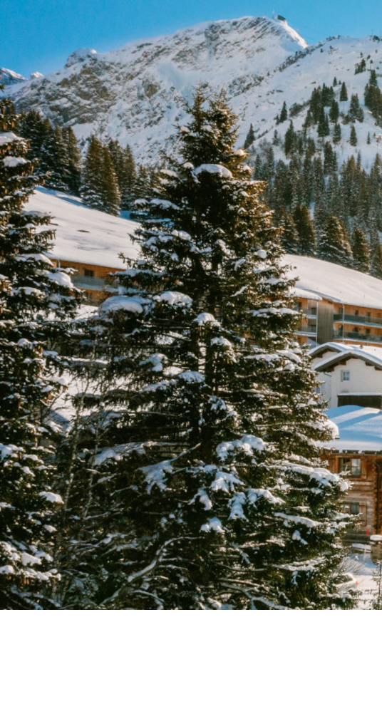 Panoramic view of Lech – The Alpine Retreat Snowy alpine landscape with sun above Hotel Severin*s in Lech, surrounded by mountains and trees.