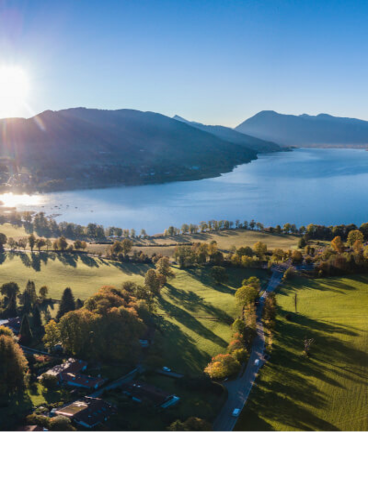 Severin*s Resort & Spa Tegernsee Luftaufnahme eines Sees mit umliegenden grünen Wiesen und Bergen im Hintergrund bei Sonnenaufgang.