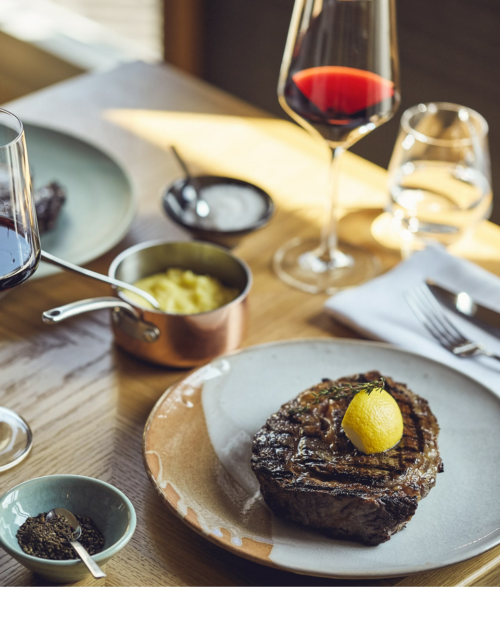 Grillroom restaurant dinner LOUIS Hotel Munich Steak with lemon, red wine glass, mashed potatoes and spices on a wooden table at the LOUIS Hotel Munich.