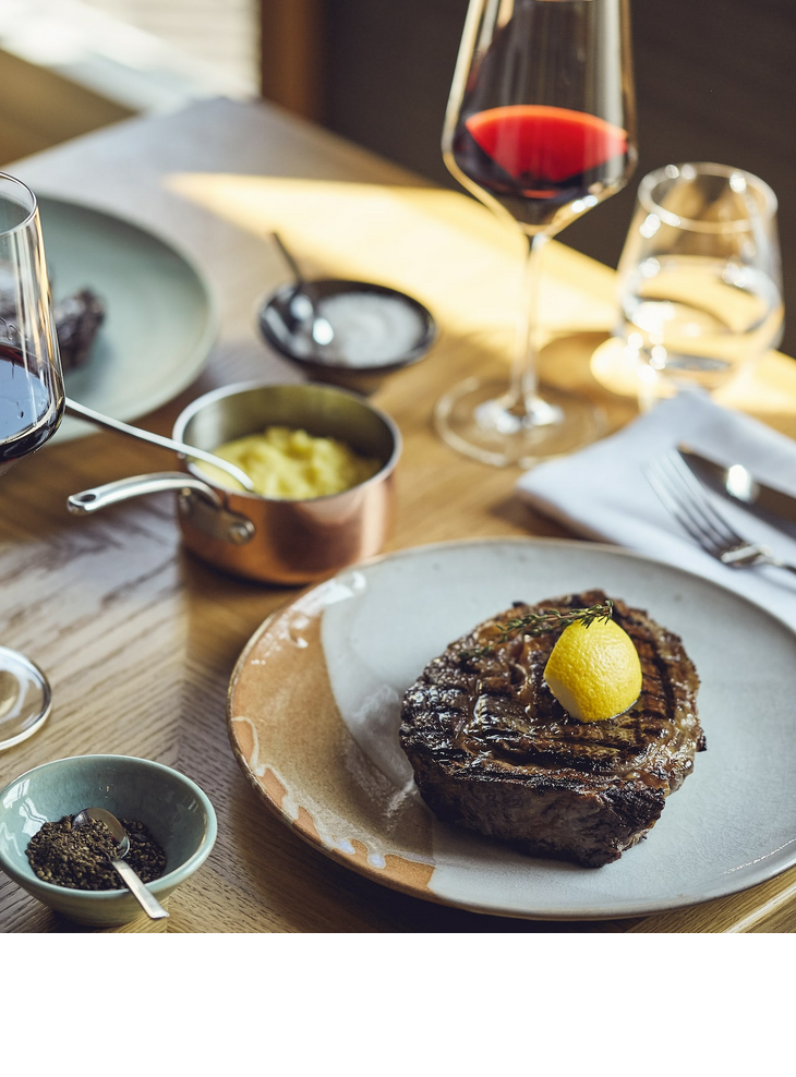 Grillroom restaurant dinner LOUIS Hotel Munich Steak with lemon, red wine glass, mashed potatoes and spices on a wooden table at the LOUIS Hotel Munich.