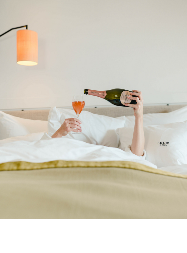 The LOUIS Hotel in Munich | Offer weekend rate Person in bed pouring champagne into a glass, surrounded by white cushions at the LOUIS Hotel Munich.